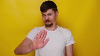 Ciddi genç adam dur hareketi gösteriyor, hoşnutsuz adam kameraya bakıyor yanlış telkin veya tekliften kaçınıyor, beyaz tişört giyiyor, sarı stüdyo arka planında kopya alanı olan izole edilmiş.