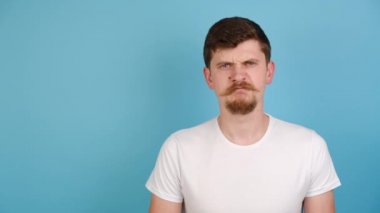 Memnuniyetsiz genç adam baş parmak hareketi gösteriyor, mutsuz milenyum adamı müşteri ürün ya da servis hakkında olumsuz geri bildirim yapıyor, beyaz tişört giyiyor, mavi arka planda kopya alanı ile izole edilmiş.