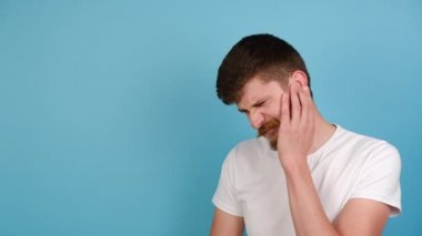 Kulak ağrısı çeken sağlıksız bir adam. Kulak tıkayan mutsuz genç bir adam, acı veren bir rahatsızlık hissediyor, kötü sesten kaçınıyor, pastel mavisi arka planda fotokopi çekilen t-shirt 'ler giyiyor.