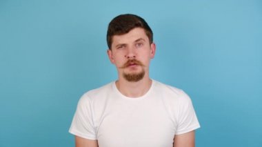 Mutsuz genç adam sıkılmış, motivasyon eksikliği, ilgisizlik, mavi stüdyo arka planında izole edilmiş tişört giyiyor. Suratı asık üzgün adam doğrudan kameraya bakıyor. İnsan duyguları kavramı