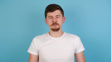 Genç adam rahatlamaya çalışıyor, evde meditasyon yapıyor, ellerini zen jestine sokuyor, gözlerini kapatıyor, beyaz tişört giyiyor, maviden izole edilmiş arka planda fotokopi alanı var. İnsanlar, rahatlama, yoga konsepti
