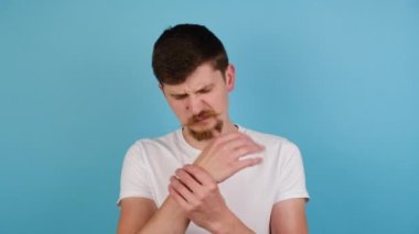 Milenyum delikanlısı romatizma iltihabından muzdarip el masajı yapıyor, bileğe dokunuyor eklem ağrıları çekiyor osteoartrit hastalığı sağlık sorunu yaşıyor, mavi stüdyo arka planında izole edilmiş.