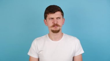 Milenyum adamı, mavi stüdyo arka planında izole edilmiş, parmaklarıyla burnunu sıkan, iğrenç bir şey kokan, iğrenç bir ifadeyle kameraya bakan, beyaz tişört giyen, kötü koku hakkında kötü hisseden.