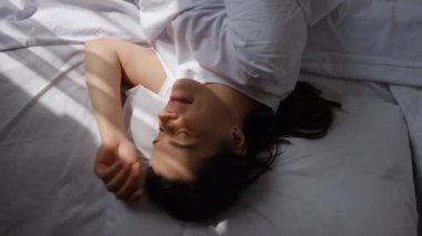 Çekici, dinlenmiş, dinlenmiş bir kadın evdeki rahat yatak odasında uyanmaktan zevk alıyor. Beyaz yatakta uzanan güzel esmer kadın sabah güneşinin altında uyanır.