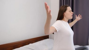 Beyaz yatakta oturup yoga yapan hamile bir kadının yan görüntüsü. Sakin, çekici, müstakbel anne kendini evinde rahat hissediyor. Rahatlama ve yoga kavramı