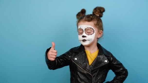 Kleines Mädchen hat kreatives buntes Gesicht, Daumen hoch Geste, zeigt Erfolgszeichen, isoliert auf blauem Hintergrund, gekleidet in Halloween-Kostüm, trägt Zuckerschädel Make-up. Todestag in Mexiko-Stadt