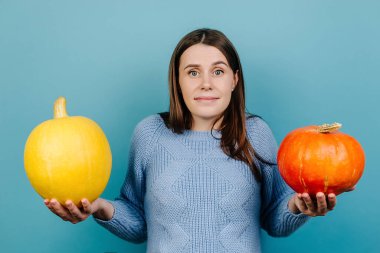 Kafası karışmış kadınların omuz silkmelerinin stüdyo fotoğrafı, belirsiz bir yüz ifadesi, hangi balkabağını seçemeyeceği, süveter giyeceği, kapalı stüdyo arka planında modeller olması. İnsanlar ve sonbahar konsepti