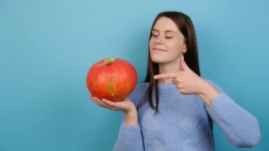 Tatlı bir kadın, olgun bir balkabağını işaret eder. Sonbaharı ve cadılar bayramını simgeler. Örgü süveter giyer. Mavi stüdyo duvarında izole edilmiş, sebzeli gülümseyen bir kız. Sağlıklı gıda ve hasat kavramı