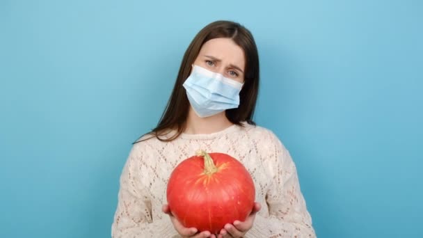 コロナウイルスCovid 19パンデミック隔離の概念の間のハロウィーン 医療用マスクを身に着けている若い女性の肖像画は 青い背景に隔離された熟した秋の野菜とオレンジカボチャ ポーズを保持します — ストック動画