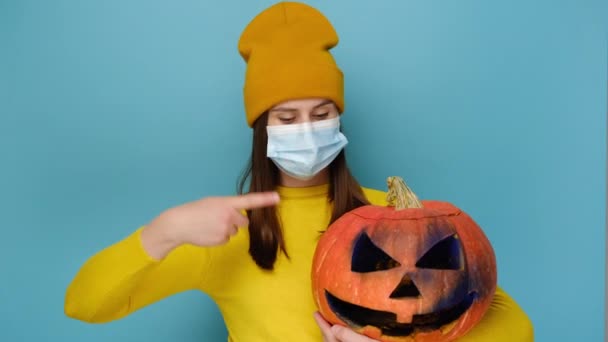 医学の顔のマスクの若い女性は 不気味なカボチャの点指は 青いスタジオの背景に隔離されたオレンジ色のセーターと帽子を身に着けています コロナウイルスCovid 19パンデミック隔離コンセプト中のハロウィーン — ストック動画