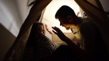 Genç baba ve şirin anaokulu kızı, Cadılar Bayramı süslemelerinin arasında karanlık çadırda oturup geceleri yatmadan önce birbirlerine ürkütücü hikayeler anlatırlar.