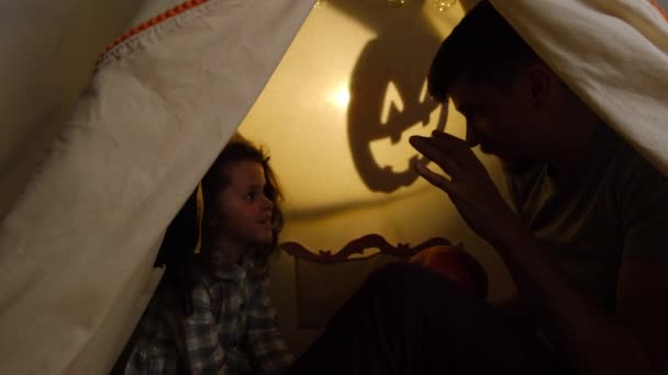 小女儿坐在黑暗的帐篷里 坐在装饰品中 向父亲讲述恐怖的故事 形似阴影笼罩的南瓜 验尸官大流行期间的万圣节 自我隔离概念 — 图库视频影像