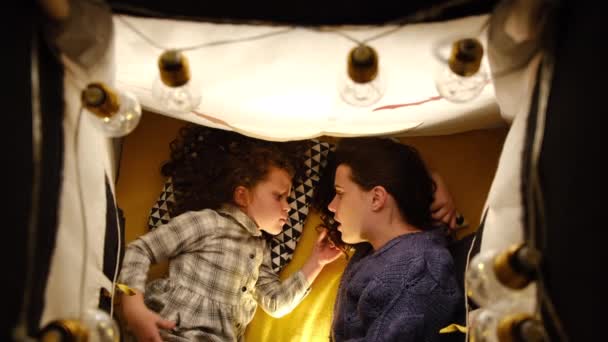 在万圣节的装饰中 年轻的母亲和小女儿在黑暗的帐篷里 以蝙蝠和花环的形式一起躺在家里 在晚上睡觉前相互讲述恐怖的故事 — 图库视频影像