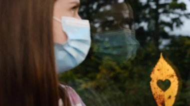 Koruyucu mavi maske takan, karantina sırasında kalp şeklinde deliği olan küçük sarı yaprağın yanında oturan üzgün, hasta bir kadının yakın plan portresi. Kavram salgını. Seçici odak