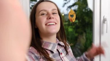 Pozitif kadın pencerenin yanında kalp şeklinde bir delikle dikiliyor. Fotoğraf makinesine el sallıyor. Kız arkadaşlarıyla sohbet ediyor.