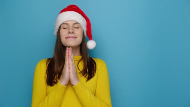 年轻女子的画像 双手交叉着祈祷 祈求一件好东西让手指交叉 身穿黄色毛衣 头戴圣诞帽 在蓝色的墙壁上摆姿势 新年快乐假日快乐 — 图库视频影像
