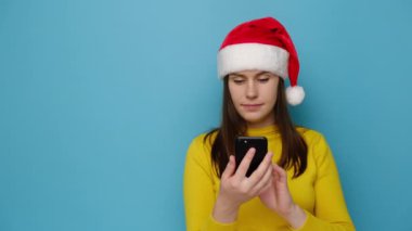 Elinde akıllı telefon tutan gülümseyen kadın, harika haberlerle mesajlaşırken mutlu hissediyor. Fotokopi alanına işaret parmağı, süveter ve Noel şapkası takıyor, mavi duvarda izole edilmiş bir şekilde işaret ediyor. Mutlu yıllar.