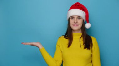 Mutlu esmer bayan avuç içini havada tutuyor, reklam içeriğiniz için ön parmağı boş, Noel şapkası ve sarı süveter giyiyor, mavi stüdyo duvarında izole edilmiş. Yeni yıl tatili