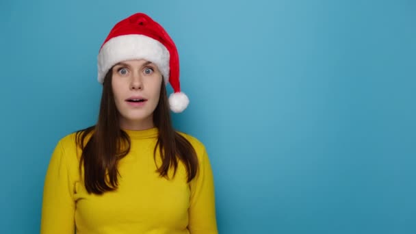 興奮した女の子の肖像画クリスマスの帽子を矢印で広告のためのスペースをコピーすることを指して身に着けている黄色のセーター 青の背景に隔離された ハッピーニューイヤーお祝いメリーホリデーコンセプト — ストック動画