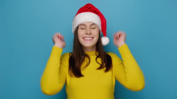 陽気な驚きの若い女性は 黄色のセーターとサンタクリスマスの帽子を着て 青いスタジオの背景に隔離された手を上げて オムグすごいと言う お正月お祝いメリーホリデー — ストック動画