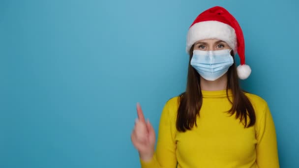 脇に手で医療マスクポイントを身に着けている素晴らしい若いサンタの女性は あなたの広告のためのコピースペースを示し 青いスタジオの壁に隔離されたクリスマスの赤い帽子とセーターを身に着けています あけましておめでとうございます — ストック動画