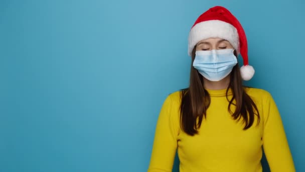 医療面マスクを身に着けている若いブルネットの女性は コピースペースと青のスタジオの背景に隔離された クリスマスの赤い帽子と黄色のセーターを身に着けている 腕を交わしてShrugs 不幸に見える — ストック動画