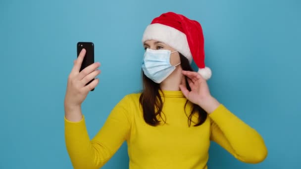 黄色のセーターの20代の若い女性手に携帯電話を保持サンタクリスマスの帽子は 青の壁に対して 自分自身をコロナウイルスから守るために保護医療用マスクを身に着けています 新常態概念 — ストック動画