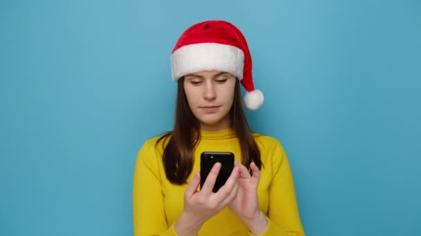 携帯電話タイプのSmsを使用して保持する衝撃的なかわいい女性は 青い背景スタジオに隔離された赤いクリスマスの帽子と黄色のセーターを身に着けています ハッピーニューイヤーお祝いメリーホリデーコンセプト — ストック動画