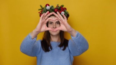 Geleneksel Noel çelengi takmış sevimli genç bir kadın. Ellerini göğsüne koyuyor. Sarı stüdyo duvarında izole edilmiş, parmakları kalp şeklinde bir hayırseverlik sembolü. Mutlu yıllar konsepti