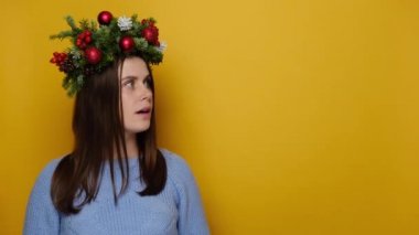 Geleneksel Noel çelengi giymiş heyecanlı genç bir kadın okla birlikte, rahat bir süveter giyiyor, sarı duvarlı arka plan stüdyosunda fotokopi alanı ile izole edilmiş. Mutlu yıllar, mutlu yıllar.