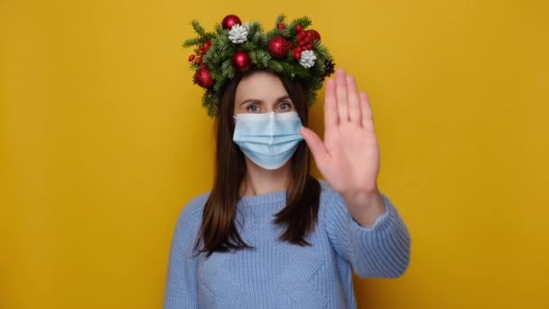 医療面のマスクで不満若い女性は タイムアウトジェスチャーを示し 停止する必要があり 黄色のスタジオの壁に隔離された手作りの花輪と青いセーターを着ています 流行性コロナウイルスとクリスマスの概念 — ストック動画