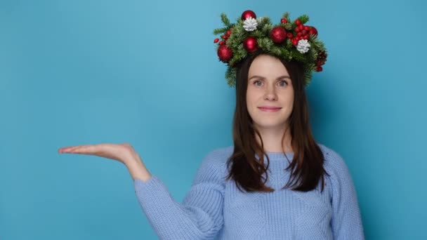 伝統的なクリスマスの花輪で楽しいかわいい若い女性は 喜んで脇を見て 居心地の良いセーターを着て 青いスタジオの背景に隔離されたコピースペースを促進します 人々と幸せな新年のコンセプト — ストック動画