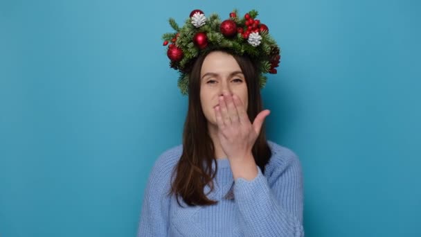 伝統的なクリスマスの花輪の美しい若い女性の肖像画は 青いスタジオの背景に隔離された立って カメラに空気のキスを送っています ハッピーニューイヤーお祝いメリーホリデーコンセプト — ストック動画