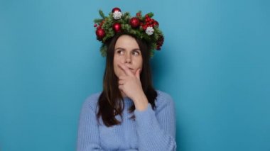 Geleneksel Noel çelengi giymiş düşünceli genç bir kadın, bir kenara bakar, parmağını çenesinde tutar, kopya alanı olan mavi stüdyo arka planında izole bir şekilde durur. Düşünceli kız bir şey hakkında rüya görüyor. Yeni yıl konsepti
