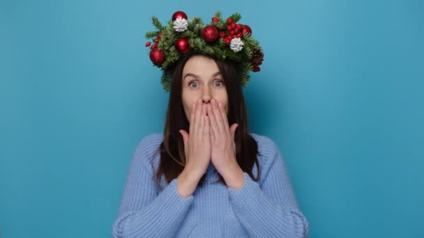 伝統的なクリスマスの花輪で幸せなショックを受けた女性は 頬に手のひらを保持し 喜びの表情をしており 居心地の良いセーターを着て 青いスタジオの背景に隔離された新年を待っています クリスマスが来る — ストック動画