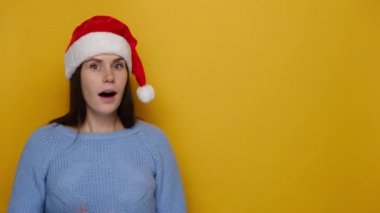 Duyarlı genç bir kadın, Noel şapkası ve rahat mavi süveter giymiş, sarı stüdyo arka planında izole edilmiş gizli bir dedikoduyu sürdüreceğine söz verdiği için fermuarlı bir jestle ağzını kaybediyor. Mutlu yıllar konsepti