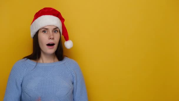 クリスマスの帽子と居心地の良い青いセーターを着て 黄色のスタジオの背景に隔離された秘密の噂を保つ約束として 動機の若い女性はジップジェスチャーで彼女の口を失う あけましておめでとうございます — ストック動画