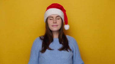 Güzel, sakin bir genç kadının portresi avuç içlerini birbirine bastırıyor Noel şapkası ve mavi süveter giyip sarı stüdyo arka planında izole edilmiş dua ediyor. Yalvarma ve mutlu yıllar konsepti