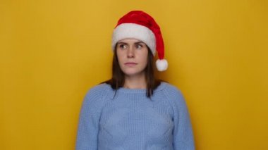 Şüpheci genç kadın sorunlu görünüyor, şaşkın duruyor, mavi kazak ve Noel kırmızı şapkası giyiyor, sarı stüdyo arka planında izole edilmiş. Mutlu yıllar, mutlu yıllar.