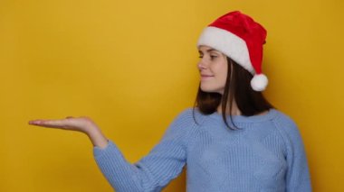 Çekici genç kadın avuç içini havada tutar, reklam için sarı arka planda işaret parmağıyla işaret eder, Noel kırmızı şapkası ve kış süveteri giyer. Mutlu yıllar, mutlu yıllar.