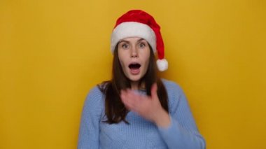 Şaşırmış genç bir kadın avucunu ağzında tutuyor, aşırı neşeli bir yüz ifadesi var, rahat bir süveter giyiyor ve Noel kırmızı şapkası takıyor, yeni yılı bekliyor, sarı stüdyo arka planında izole edilmiş. Xmas geliyor.