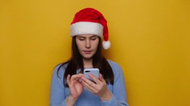 Tatmin olmuş genç bir kız cep telefonuna kısa mesaj yazıyor, çevrimiçi iletişimden hoşlanıyor, geri bildirim yapıyor, Noel kırmızı şapkası ve kışlık kazak giyiyor, sarı stüdyo duvarına izole edilmiş. Xmas, kutlama.