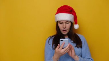 Şok olmuş bir kadın cep telefonu kullanıyor, SMS yazıyor ve fotokopi alanını işaret ediyor, kırmızı Noel şapkası ve rahat süveter giymiş, sarı duvarda izole edilmiş. Yeni yılı kutluyoruz. Mutlu tatiller.