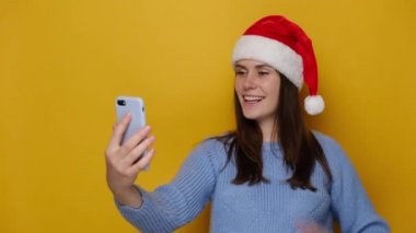 Noel Baba şapkalı genç bir kadın cep telefonu kullanıyor, selfie video konferansı yapıyor mavi rahat bir süveter giyiyor, sarı duvarda fotokopi alanı ile izole edilmiş. Mutlu yıllar, mutlu noeller.