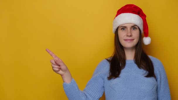クリスマスの帽子の魅力的な千年紀の若い女性は 承認されたカメラの魅力的な笑顔を見て 黄色のスタジオの背景に隔離された青いセーターを着ています あけましておめでとうございます — ストック動画