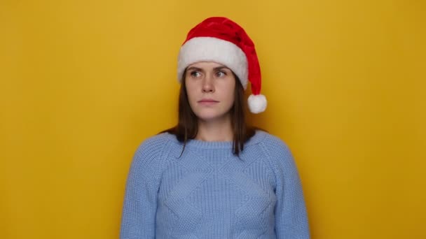 疑わしい若い女性はさておき 困惑スタンド 青い居心地の良いセーターやクリスマスの赤い帽子を着て 黄色のスタジオの背景に隔離された問題に見える お正月お祝いメリーホリデー — ストック動画