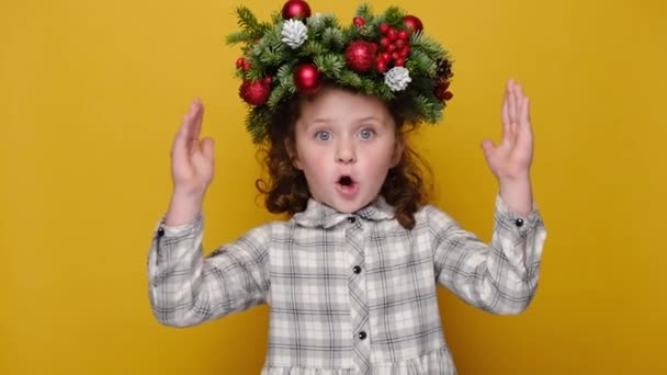 手で大きなサイズのジェスチャーを示すかわいい子供の女の子を驚かせ 感銘を受けた子供は 黄色のスタジオの背景に隔離された手作りの花輪を身に着けています メリークリスマスのコンセプト — ストック動画