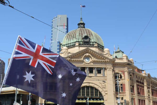 МЕЛЬБУРН, Австрия - 26 ЯНВАРЯ 2019: Австралийский флаг перед железнодорожным вокзалом Iconic Flinders Street в Мельбурне

