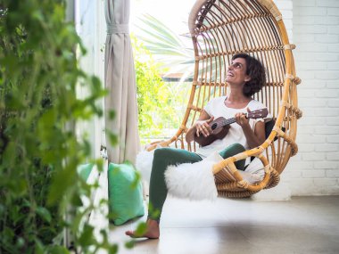 Ukulele oynayan genç bir kadın salıncakta oturuyor, rahatlıyor ve tatil konsepti yapıyor.