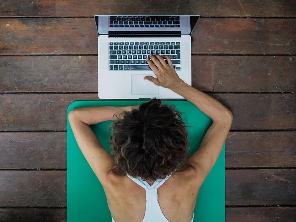 Evde dizüstü bilgisayar kullanan mutlu bir kadın yoga minderinde yatıyor.
